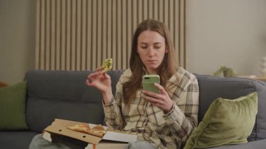 Samimi oturma odasında pizza yerken Smartphone 'a konsantre olan bir kadın. Rahatlamış Kadın, Ekrana Odaklanmış, Pizzanın Zevkini Çıkartıyor, Rahat Bir şekilde Oturuyor