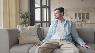 Öğrenci koltukta oturuyor, kontrolörü eline alıyor ve bir video oyunu seansı başlatıyor. Genç erkek evde eğleniyor, Parlak Modern Salon Ortamı 'nda Oyun Faaliyetleri Yapıyor.