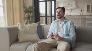 Parlak, modern oturma odasında video oyununun tadını çıkaran, gülümseyen ve rahatlatan bir adam. Rahatlamış Oyuncu, kontrolörle rahat bir şekilde oturuyor, Gün Işığında rahat bir şekilde ev içlerinde eğleniyor.