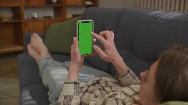 Kadın rahat odasında yeşil ekranlı akıllı telefondan kanepeye uzanıyor. Uygulama Modeli, İçerik Görünümü veya Sosyal Medya Şablonu Kullanımı için Boş Ekranlı Sıradan Boş Zaman Anı