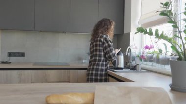 Kıvırcık Kadın alışverişten sonra Bright Cozy Kitchen 'daki pencerenin yanında sıcak içecek hazırlıyor. Genç bir kadın, modern iç mekandaki bakkaliyeleri açtıktan sonra çayla kaplanırken evde rahat bir an.