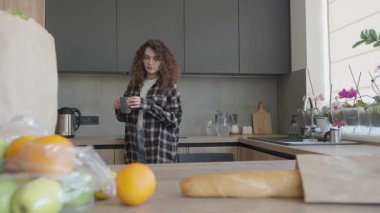 Kıvırcık Kadın paketleri açtıktan sonra Bright Kitchen 'da sıcak içeceğin tadını çıkarıyor. Alışverişten sonra evde çay ya da kahve içen rahat bir kadın, yakın zamanda marketin içinde ayakta ve rahat bir şekilde oturuyor.