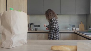 Bakkal alışverişinden sonra sıcak mutfakta çay demlemek için çay poşetine uzanan bir kadın. Doğal ışık, sıcak içeceği hazırlarken, evdeki huzur verici alışveriş sonrası rutinini yakalıyor.