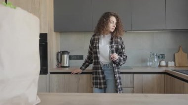 Kıvırcık Kadın Bardağı Tutuyor, Modern Parlak Mutfakta Çay Yapmaya Hazırlanıyor. Minimalist iç mekanda samimi sabah ritüeli, marketten sonra sıcak içecekle huzurlu anın tadını çıkaran kadın.