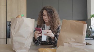 Kıvırcık Kadın, marketteki eşyaları akıllı telefonu evde kullanırken makbuzla karşılaştırıyor. Ev Bütçesinin Otantik Anı ve Sıcak Mutfakta Akıllı Alışveriş. Yüksek kalite 4k görüntü