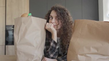 Market poşetlerinin arasında oturan kadın, Parlak Mutfak 'ta paketleri açmadan önce duraklıyor. Rahat genç bayan alışverişe çıktıktan sonra, rahat modern iç mekanlarda taze yiyeceklerle çevrili.