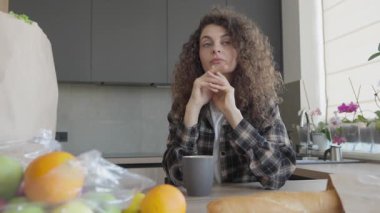 Uzun bir günden sonra mutfakta elinde kahveyle oturan dalgın kadın. Yakın çevredeki taze bakkaliyelerle Parlak Konforlu İçten Yansımanın Huzurlu Anı, Yaşam Tarzı ve Sağlık Teması İçin İdeal