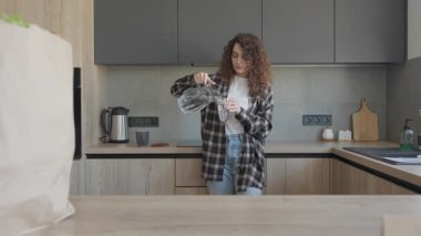 Alışveriş paketlerini açtıktan sonra modern mutfakta su içen rahat kadın. Genç bayan alışveriş yaptıktan sonra dinlenmenin keyfini çıkarıyor, arka planda market poşetleriyle şık, parlak bir mutfakta duruyor.