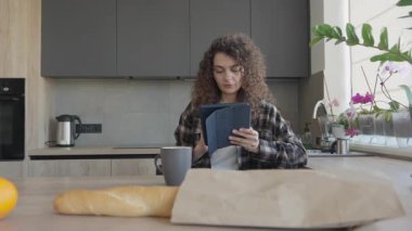 Mutfakta yiyecek ve kupalarla otururken tablette kitap okuyan bir kadın. Flanel tişörtlü kıvırcık kadın mutfak masasında dinleniyor, odaklanmış ifade ile içeriğine bakıyor.