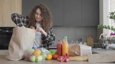 Gülümseyen kadın paketleri açıyor ve sıcak modern mutfakta taze baget yiyor. Alışveriş yaptıktan sonra etrafı meyveler, meyve suları ve sebzelerle çevrili neşeli kadın organizasyonu.