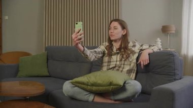 Kanepede bacak bacak bacak üstüne atarak oturan bir kadın videoda gülüyor ve konuşuyor. İçerde dostça sohbet, rahat atmosfer, Mutlu İfade, Samimi Bağlantı Anı ve Gerçek Duygu Ekranda