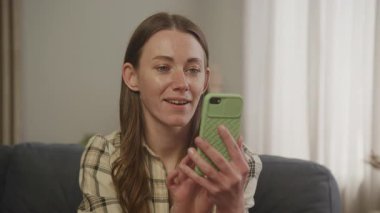 Evde video görüşmesi yaparken el sallayan olumlu bir kadın koltukta rahatça oturuyor. Dostça İletişim Anı, Rahat Kapalı Salon Atmosferi, Gerçek Bağlantı, Uzaktan Sohbet, Modern Yaşam Tarzı