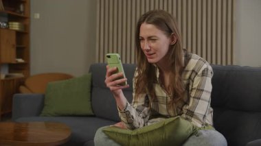 Mutlu Kadın, evdeki kanepede eğlenirken yüksek sesle gülüyor. Etkileyici Kadın Neşeli Sohbetten Hoşlanıyor, Rahat Oturma Odasında Rahatlıyor, Akıllı Telefonu Gerçek Duygu ve Enerjiyle Tutuyor