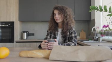 Dolu bir günden sonra elinde bardak tutan kadın mutfak penceresinden dışarı bakıyor. Huzurlu bir an, evdeki günlük hayatı yakalıyor, rahatlama, rahatlık, farkındalık, ev rutini ve kişisel bakım gibi kavramlar için mükemmel.