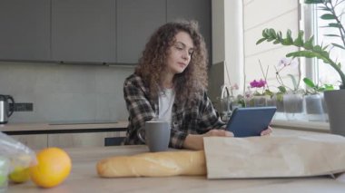 Bardaktan içen ve mutfak masasında erzakla tablet okuyan bir kadın. Huzurlu Anlar Modern Yaşam Tarzı, Ev Konforu, ve Parlak Dijital Rutin, Yüksek Kaliteli 4k Görüntü