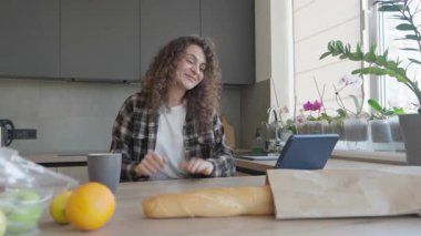 Mutfakta görüntülü konuşma yapan gülümseyen kadın, olumlu haberlere tepki veriyor. Arkadaş Canlısı İletişim, Modern Ev İçi Neşeli Kadın, Alışveriş Sonrası Çevrimiçi Bağlantının Tadını Çıkartıyor