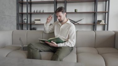 Koltukta oturan, elinde kalemle oturup not defterlerini okuyan bir adam. Sakin Atmosfer, Yaratıcı Beyin Fırtınası Anı, Uzaktan Çalışma, Serbest Çalışmalar, Ev Eğitimi gibi kavramlar için ideal