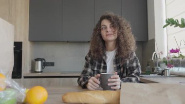 Alışveriş yaptıktan sonra kupayla rahat bir mutfakta oturan dalgın kadın. Huzurlu Düşünce Anı, Günlük Yaşamı Yakalama, Ev İçi Rahatlama, Sıcak Işık, Yaşam Tarzı ve Ev Temaları İçin İdeal