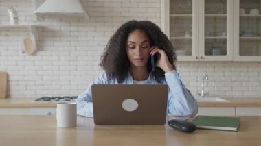 Bright Kitchen Masası 'nda telefonla konuşurken dizüstü bilgisayarda çalışan genç bir kadın. Uzaktan Uzaktan Uzaktan Uzaktan Profesyonel Çoklu Görev Odaklanma, Fikirleri İletişim ve Modern Konforlu Evdeki Çalışmaları Yönetme