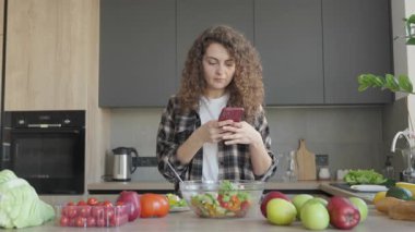 Parlak modern mutfakta salata yiyen ve akıllı telefonda yazan bir kadın. Sağlıklı Yaşam Tarzı, Evde Çoklu Görev, Dijital Rutin, Dengeli Beslenme, Sıcak Atmosfer, Günlük Gündelik Günler. Yüksek kalite 4k görüntü