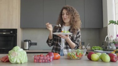 Kıvırcık saçlı kadın evdeki rahat modern mutfakta taze salatanın tadını çıkarıyor. Sağlıklı Beslenme Konsepti, Rahat Ev Atmosferi, Organik Yaşam Tarzı, Doğal Malzemeler, Taze Gıda Hazırlama