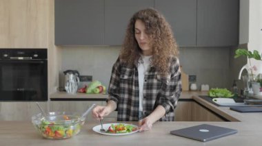 Modern mutfakta kadın salata yiyor ve laptoptan e-postaları kontrol ediyor. Üretken Sabah Rutini, Sağlıklı Yaşam tarzı, Uzaktan Çalışma Konsepti, Konforlu Atmosfer, Dijital Yaşam Dengesi, Taze Gıda Sahnesi