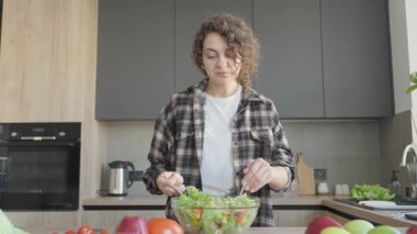 Kıvırcık saçlı kadın modern mutfakta taze salata karıştırıyor ve tadıyor. Evde sağlıklı beslenme, sebzelerle yemek pişirme, günlük sağlık rutini, doğal bileşenler, ev yapımı yemeklerden keyif alma.