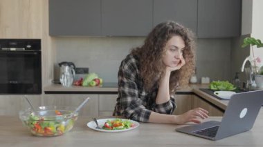 Konforlu mutfakta yemek yerken dizüstü bilgisayarda çalışan odaklanmış bir kadın. Üretken Uzaktan Çalışma Anı, Dengeli Yaşam Tarzı, Günlük Ev Ayarları, Besleyici Gıda, Dijital Düzenleme