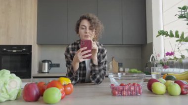 Bright Kitchen 'da taze meyvelerle elma yiyen ve akıllı telefonlara bakan bir kadın. Günlük Sabah Anı, Sağlıklı Yaşam Tarzı, Doğal Işık, Rahatlamış Ruh hali, Dijital Rutin, Organik Besin. 