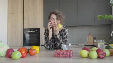 Etrafı meyvelerle çevrili, elmalı bir mutfakta telefonla konuşan genç bir kadın. Günlük Sabah Anı, Sağlıklı Yaşam Tarzı, Doğal Işık, Ev Atmosferi, Neşeli Konuşma, Gerçek Hayat Sahnesi