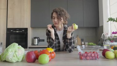 Elma tutan kadın, masadaki malzemelere bakarken yemek pişirme fikri ediniyor. Yaratıcı Mutfak Anı, Taze İlham, Sağlıklı Yaşam Tarzı, Doğal Gıda, Gelişigüzel Ev Ayarları, Gerçek Hayat Sahnesi