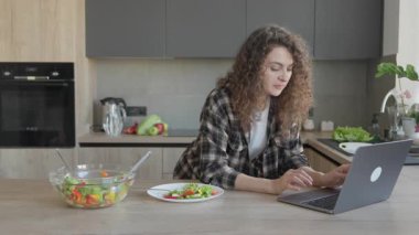 Modern mutfakta öğle yemeği sırasında dizüstü bilgisayara bakarken eline yaslanan kadın. Günlük iş anı, sağlıklı yaşam tarzı, rahat iç mekan, dijital rutin, rahat ev titreşimleri, dengeli gün sahnesi