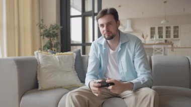 Öğrenci kaybettikten sonra oyun kontrolörünü düşürür, evdeki koltukta oturur. Modern Parlak Oturma Odası 'nda Oyun Yenilgisinden Sonra Hayal kırıklığına uğrayan Genç Adam. Yüksek kalite 4K görüntü.