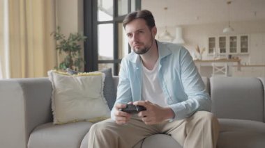 Adam kasıtlı olarak modern apartmandaki koltukta konsol oyununda yarışıyor. Evde Odaklanmış Oyun Deneyimi, Duygu, Yaşam Tarzı ve Modern İç Ambiyansın Mükemmel Bir Tanımı