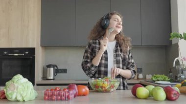 Mutfakta kaşıkla salata karıştırırken neşeyle kulaklık takmak. Mutlu Genç Kadın Sağlıklı Yemekler Pişiriyor, Modern Ev 'de pozitif titreşimli Kaşıklara Şarkı Söylüyor. Yüksek kalite 4k görüntü