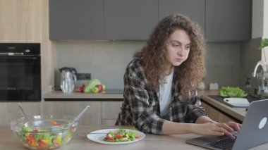 Mutfak tezgahında dizüstü bilgisayarla çalışırken sağlıklı salata yemek. Dengeli Diyet, Uzaktan Çalışma Esnekliği ve Konforlu Ev Mutfağı Konsepti