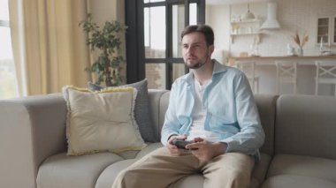 Gülümseyen Adam evdeki koltukta video oyunu oynarken zaferi kutluyor. Neşeli Oyuncu, Parlak Modern Oturma Odasında Keyifli Başarı, Haftasonu Eğlencesi. Yüksek kalite 4k görüntü