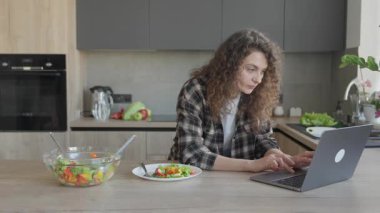 Mutfakta dizüstü bilgisayarla çalışırken tezgahta taze salata yiyorum. Uzaktan Çalışmanın Konsepti, Sağlıklı Yaşam Tarzı, Ev Ofisi Rutini, Yemek molaları, Ev Arası Üretim Ortamı İçin Mükemmel Görsellik