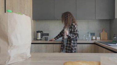 Kıvırcık Kadın Market alışverişinden sonra mutfakta telefona bakıyor. Doğal Parlak İçi Açık Masa Üzerinde Masa Üzerinde Akıllı Telefonla Bekleyen Sıradan Kadın Karakteri