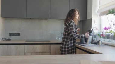 Parlak mutfak penceresinden bakarken Kıvırcık Kadın çaydanlığı suyla dolduruyor. Huzurlu Sabah Anı Günlük Günlük rutin, Ev Yaşam Tarzı, Samimi Mutfak Atmosferi. Yüksek kalite 4k görüntü