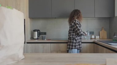 Kıvırcık kadın alışveriş yaptıktan sonra sıcak mutfağında su içiyor. Yiyecek paketlerini açtıktan sonra ferahlatıcı susuzluk anı, modern modern ev ortamında sağlıklı yaşam tarzını kucaklama