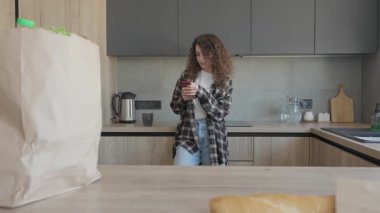 Kadın, rahat modern mutfakta dururken telefon mesajı okuyor. Flanel tişörtlü sıradan bir kadın market poşetleriyle tezgahın yanında duruyor, Sakin Ev Atmosferindeki Bildirimleri kontrol ediyor.