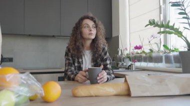 Mutfak masasında sıcak bir şeyler yudumlayan bir kadın. Rahat bir iç mekanda huzurlu bir sabah rutini. Alışveriş yaptıktan sonra yiyecek, konfor ve kişisel bakım ile ev hayatı.