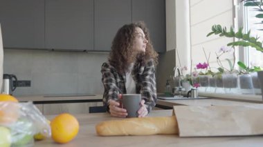 Sıcak içecek içen ve güneşli mutfak penceresinden dışarı bakan genç bir kadın. Sıcak Işıklı Huzurlu Sabah Rutini, Taze Bakkaliye, Rahatlama, Farkındalık ve Ev Hayatı