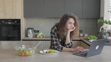 Sıcak mutfakta yemek yerken dizüstü bilgisayarda çalışan bir kadın. Odaklanmış Profesyonel Dengeleyici Uzaktan Çalışmalar ve Parlak Ev Ayarlarında Besleyici Beslenme. Sağlık ve Yaşam Tarzı İçin İdeal Manzara