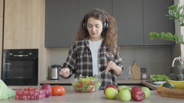 Samimi mutfak sahnesinde müzik dinlerken taze salata yemek. Sağlıklı ev yapımı yemeklerin keyfini çıkaran neşeli kulaklık takan, meyvelerle çevrili, Parlak Ev Mutfağı Atmosferinde sebzeler