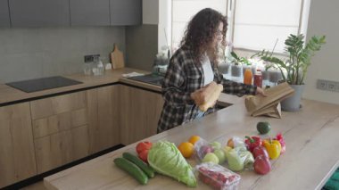 Market koşusundan sonra taze baget ve sebzeleri mutfak tezgahına yerleştiriyorum. Neşeli Kıvırcık Kadın, içi organik meyve, sebze ve Parlak Ev Mutfağında Ekmek dolu market poşetlerini açıyor.