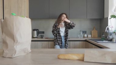 Alışverişten sonra sıcak modern mutfakta su içen bir kadın. Günlük yaşam tarzı: Hidrasyon ve Zaman Farkındalığı, Rahatlamış Ruh Haliyle Ilık Doğal Işıkta Çekilen