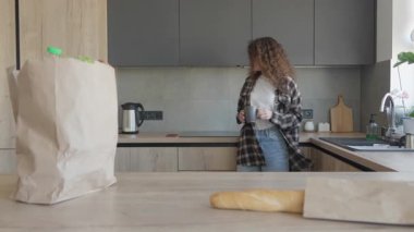 Kıvırcık Kadın, sıcak, modern mutfakta alışveriş yaptıktan sonra çay yapıyor. Rahatlamış Kadın Sıcak İçeceğe Hazırlanıyor. Etrafı yeni alınmış yiyeceklerle çevrili halde duruyor. Yüksek kalite 4k görüntü