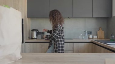 Esmer kadın çay yapıyor ve kullanılmış çay poşetini çöp kutusuna atıyor. Bakkal alışverişinden sonra sıcak içeceği hazırlayan genç bir kadının günlük hayatı ve sürdürülebilirlik rutinini gösteren günlük mutfak anı.